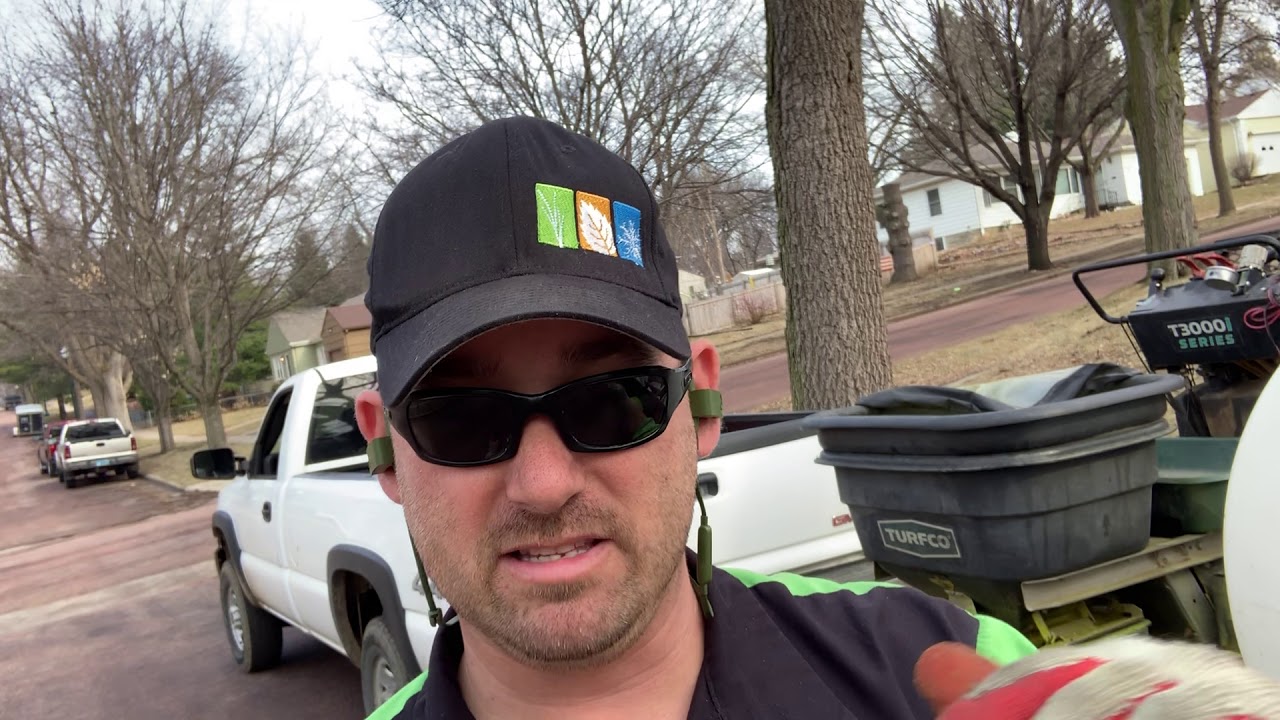 Man wearing sunglasses and a cap with a logo, discussing lawn care techniques with a focus on pre-emergent weed control, with trucks and equipment visible in the background.
