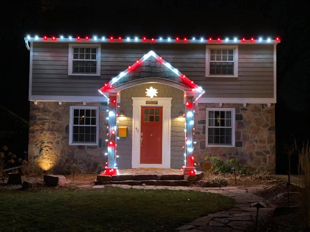 Holiday and permanent lighting adorning a charming home, featuring red and white lights outlining the roof and door, creating a festive atmosphere.