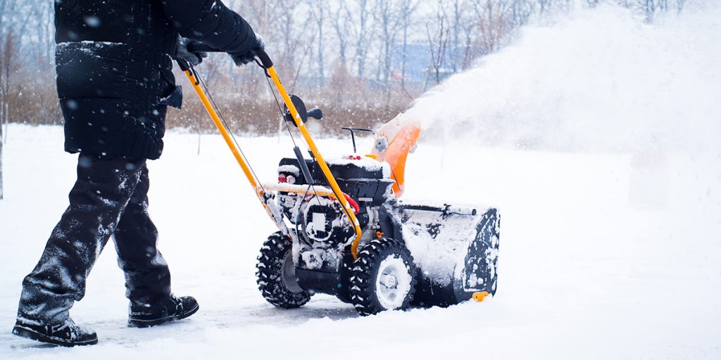 Person using a snow blower to clear snow from a residential driveway, emphasizing snow removal services in winter conditions.