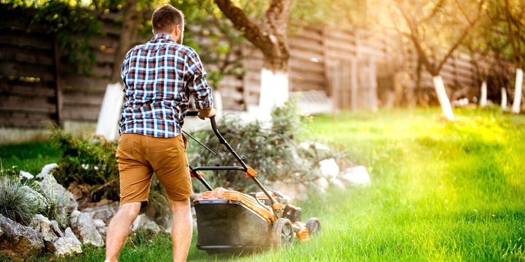 Man mowing a lush green lawn with a push mower in a residential yard, highlighting DIY lawn care efforts.